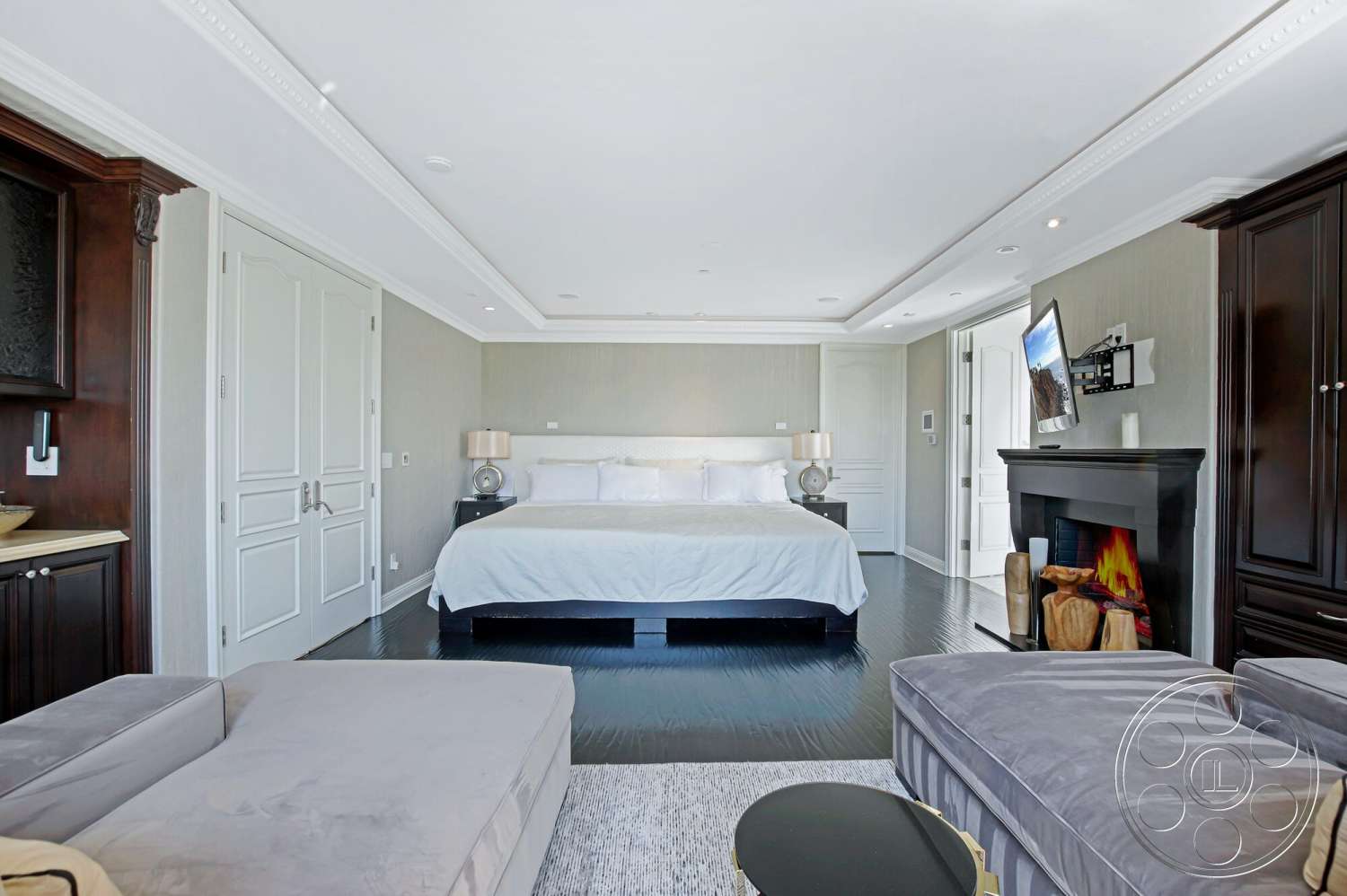 Medit 170 - dark wood cabinetry, wall-mounted television, double doors, bright natural lighting, white bedding, king size bed, modern lounge chair, black stone fireplace, hardwood bedroom floors, textured wallpaper walls, color-coordinated lamps, molding ceiling details