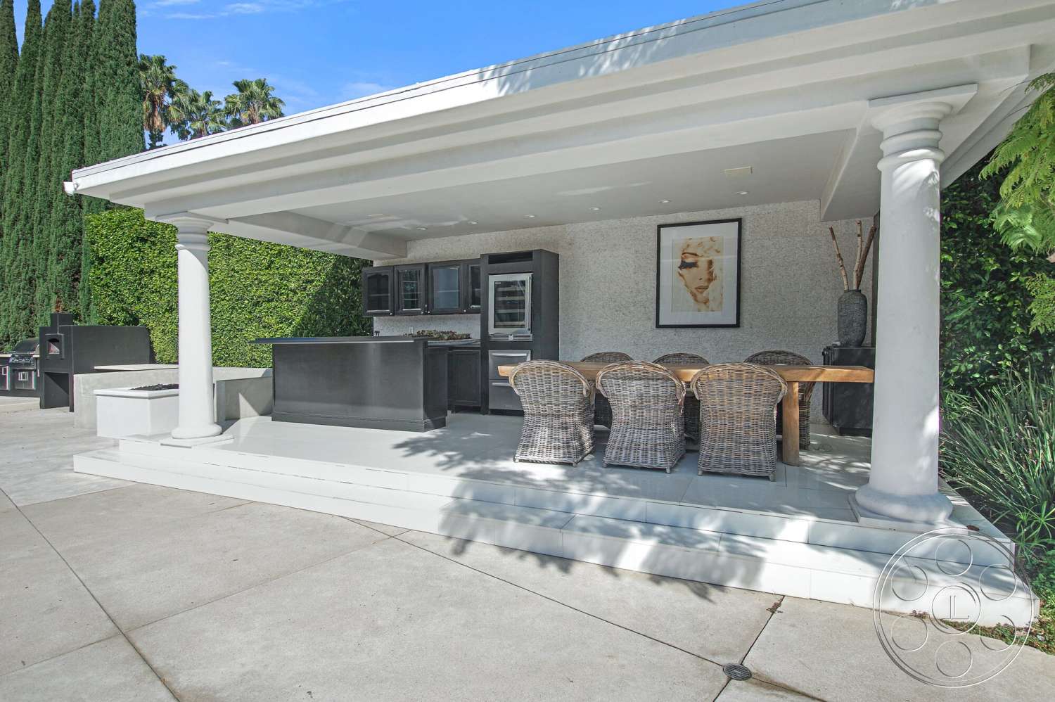 Medit 170 - exterior, dark cabinetry, outdoor kitchen area, large rectangular dining table, concrete patio flooring, stainless steel kitchen appliances, stucco wall treatment, modern outdoor living space, smooth concrete steps, woven rattan dining chairs, white painted ceiling beams, column-supported roof structure, landscaped privacy hedge