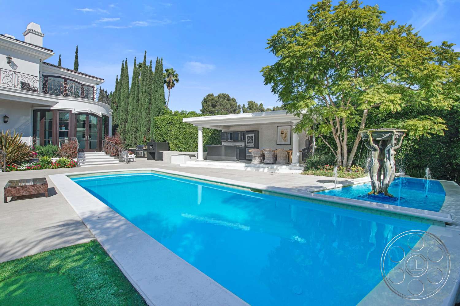 Medit 170 - exterior, green grass, white stucco walls, lush landscaping, covered patio, modern architectural style, tall cypress trees, concrete pool deck, rectangular pool, stone steps, outdoor kitchen area, bright blue sky, decorative metal railing, wooden outdoor seating, pool fountain feature, metal garden sculpture