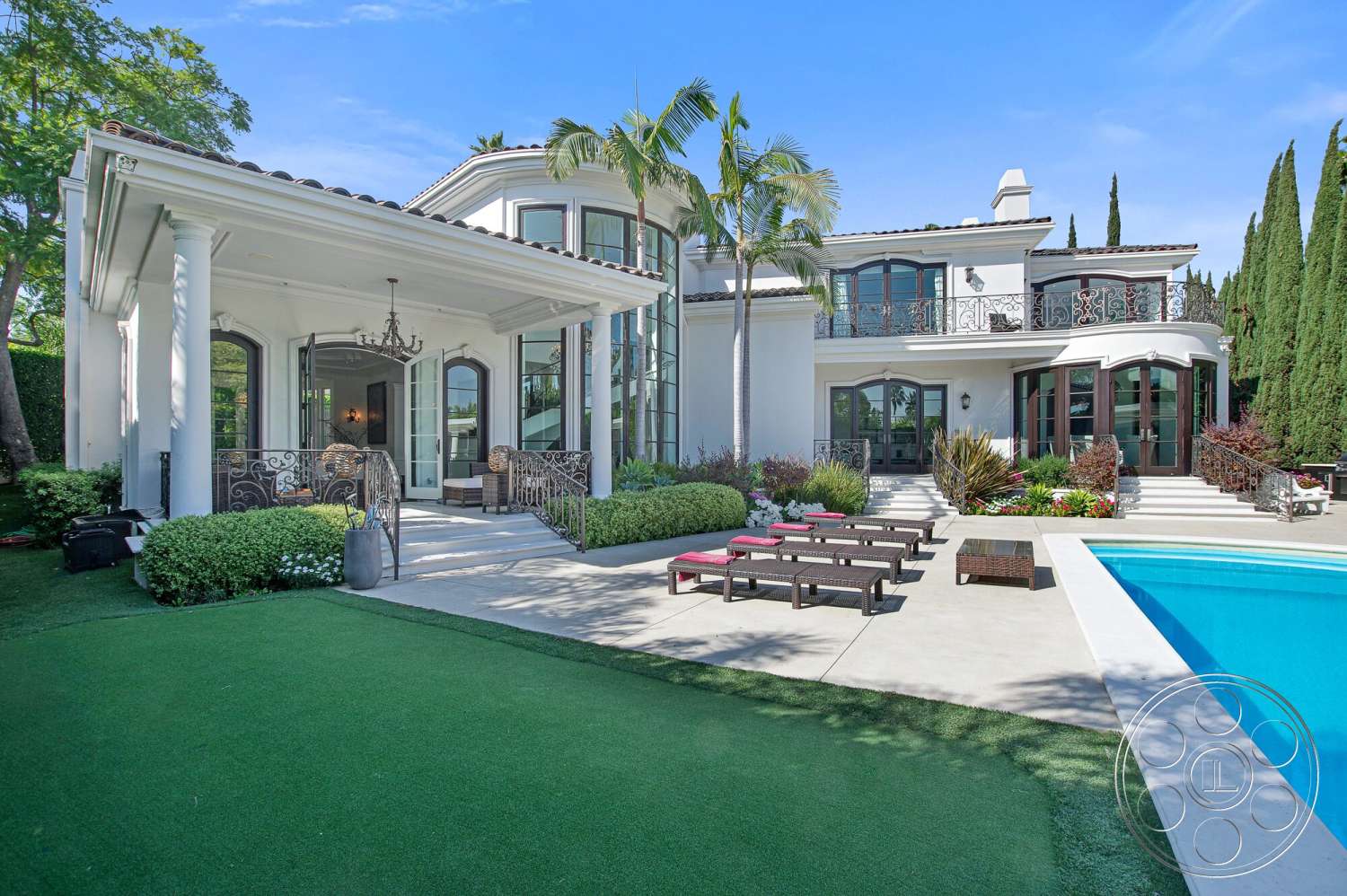 Medit 170 - exterior, rectangular swimming pool, stucco walls, tile roof, large floor-to-ceiling windows, smooth concrete pool deck, landscaped garden area, outdoor lounge seating, curved patio, modern Mediterranean architectural style, decorative wrought iron railings, covered porch area, lawn area with artificial grass, paved walkway steps, tall pine trees in backyard