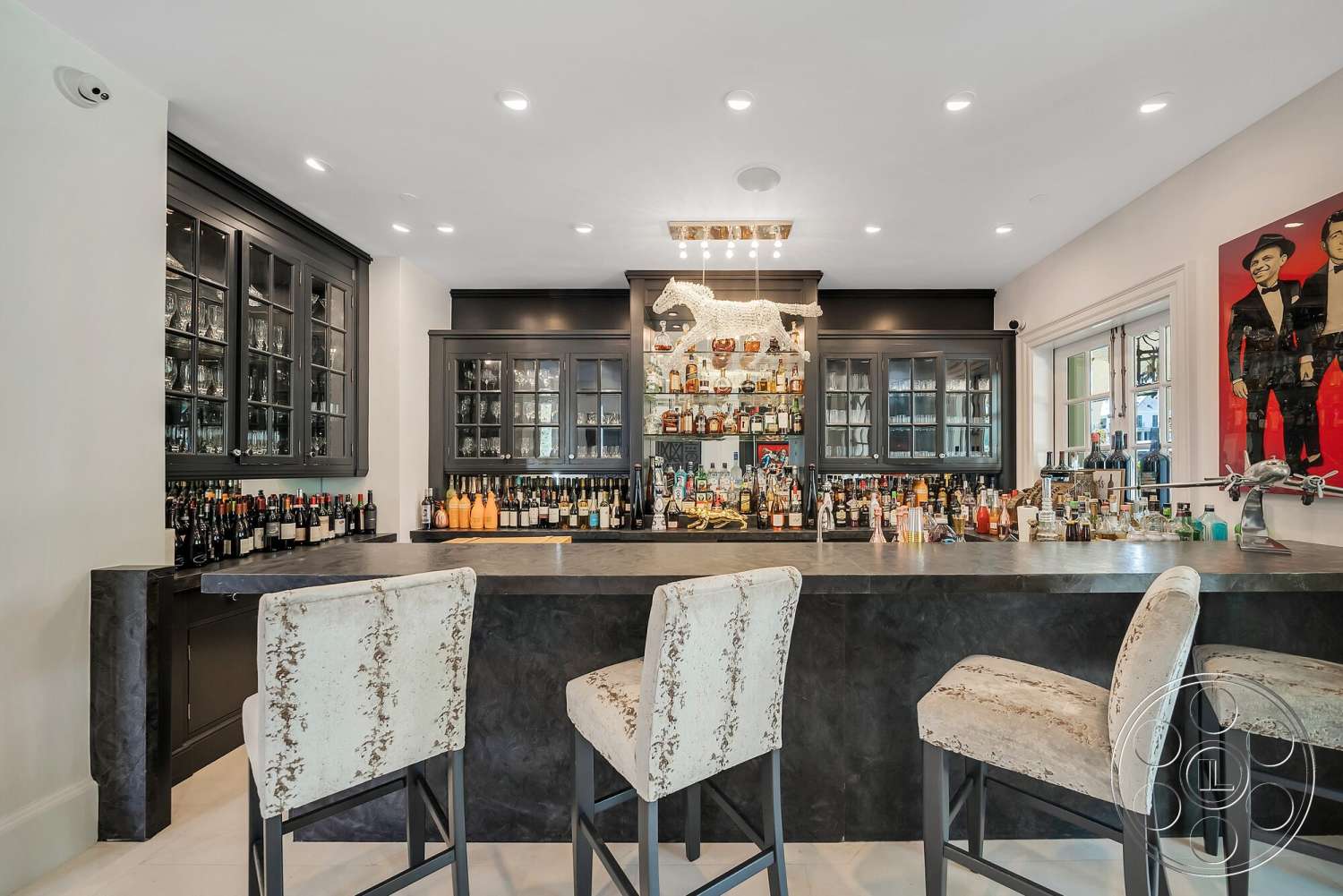Medit 171 - colorful wall art, recessed ceiling lights, interior bar area, white tile flooring, black cabinetry with glass doors, polished concrete bar countertop, upholstered bar stools with floral pattern, woven decorative art piece, bottled liquor display, luxurious drink preparation space