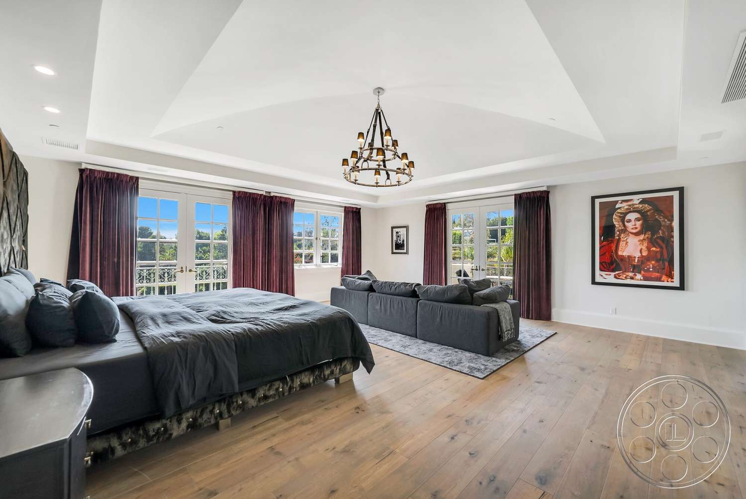 Medit 171 - open space layout, floor-to-ceiling windows, vaulted ceiling, master bedroom, gray area rug, black bed frame, hardwood bedroom floors, dark wood window frames, white painted walls, framed artwork on wall, chandelier lighting fixture, plush grey sectional sofa, sheer burgundy curtains