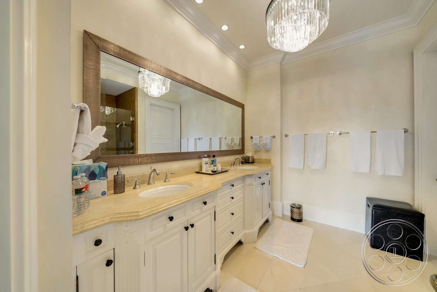 Medit 171 - exterior, glass shower enclosure, textured plaster walls, cream tile flooring, chrome faucet fixtures, built-in bathroom storage, white cabinetry bathroom vanity, cream marble countertop bathroom sink, large framed mirror above vanity, modern crystal chandelier ceiling light, white plush bath mats, towel rack with white towels