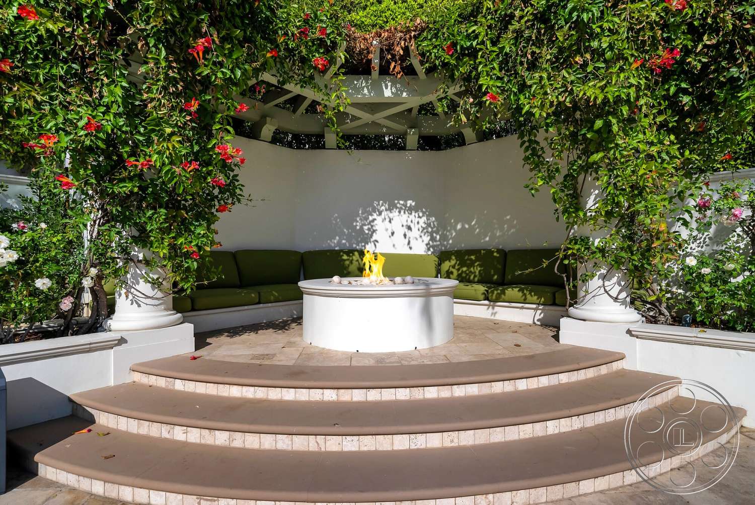Medit 171 - white stucco walls, circular fire pit, interior seating area, white decorative columns, natural stone patio flooring, modern outdoor design, curved stone steps, green upholstered outdoor sofa, lush flowering vines and greenery, outdoor shaded retreat