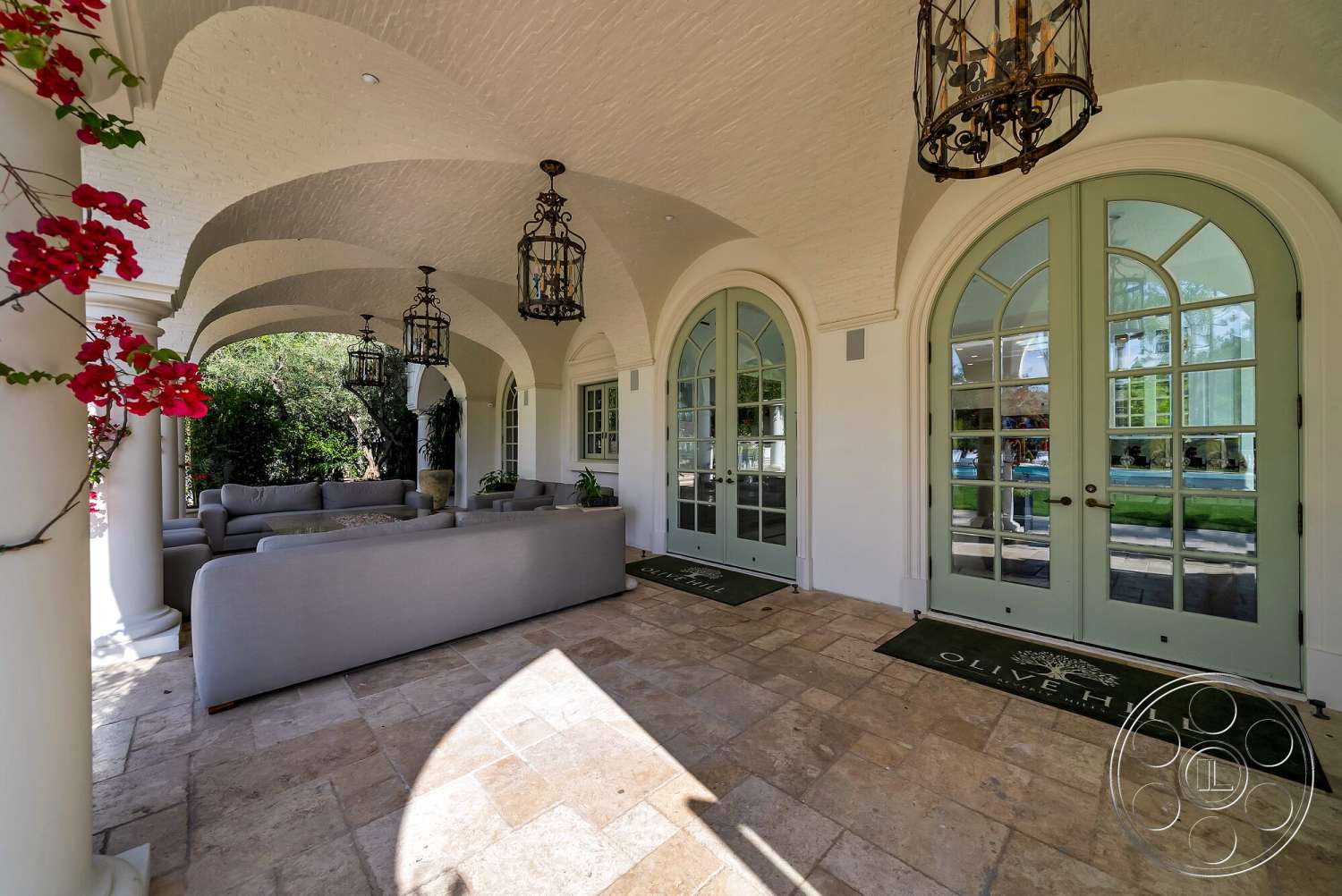 Medit 171 - exterior, potted plants, textured plaster ceiling, arched doorways, green double doors, covered porch area, hanging lantern light fixtures, stone floor patio, soft grey outdoor sectional sofa, red bougainvillea