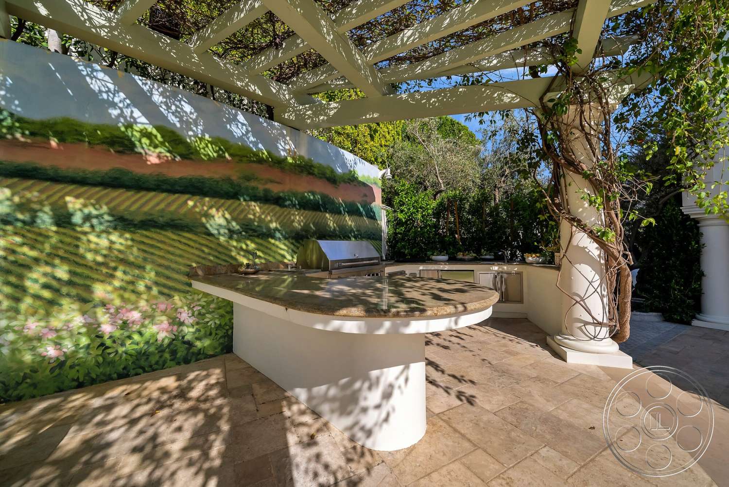 Medit 171 - exterior, wood pergola structure, lush greenery setting, stone outdoor kitchen countertop, travertine patio flooring, plastered wall with vineyard mural, round outdoor kitchen bar, decorative column