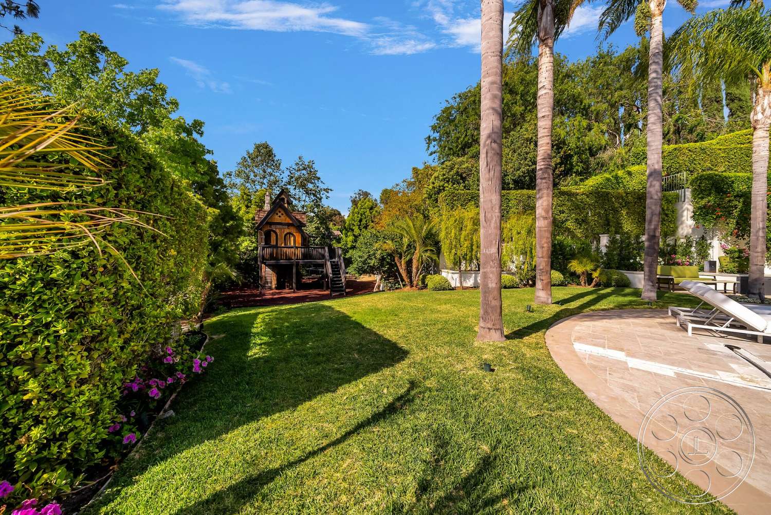 Medit 171 - exterior, palm trees, lush green lawn, flowering shrubs, tropical landscaping, sunny outdoor setting, hedge border, wooden playhouse structure, curved stone patio, partial shade from trees