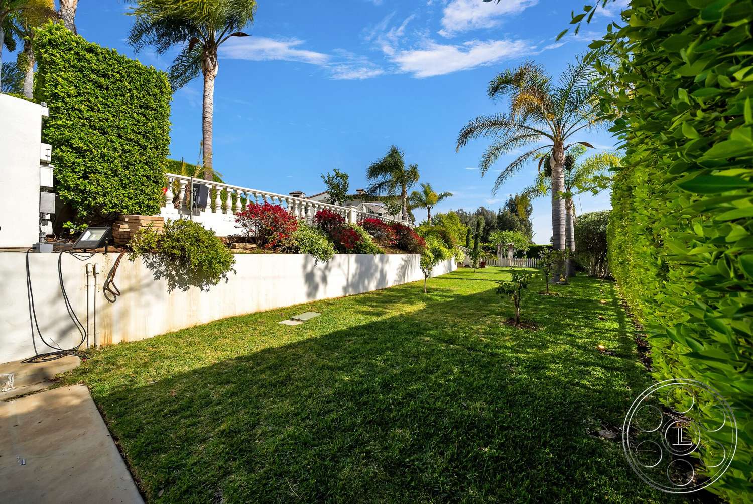 Medit 171 - exterior, landscaped garden, lush green grass, flowering shrubs, clear blue sky, tropical palm trees, colorful flower beds, concrete garden wall, white balustrade, hedged privacy fence, patio pathway, scenic backyard view