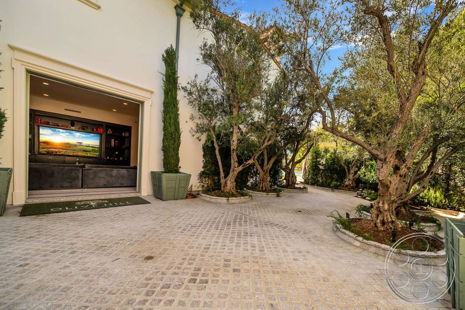 Medit 171 - stone driveway, paved stone flooring, exposed wood ceiling beams, lush garden setting, rectangular driveway, modern garage, tall olive trees, framed view of outdoor greenery, cream-colored exterior walls, built-in cabinetry with entertainment center