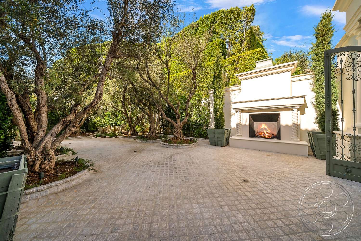 Medit 171 - exterior, lush greenery, concrete wall, metal gate, outdoor fireplace, decorative planters, landscaped garden area, mature olive trees, rectangular patio, paver stone patio flooring