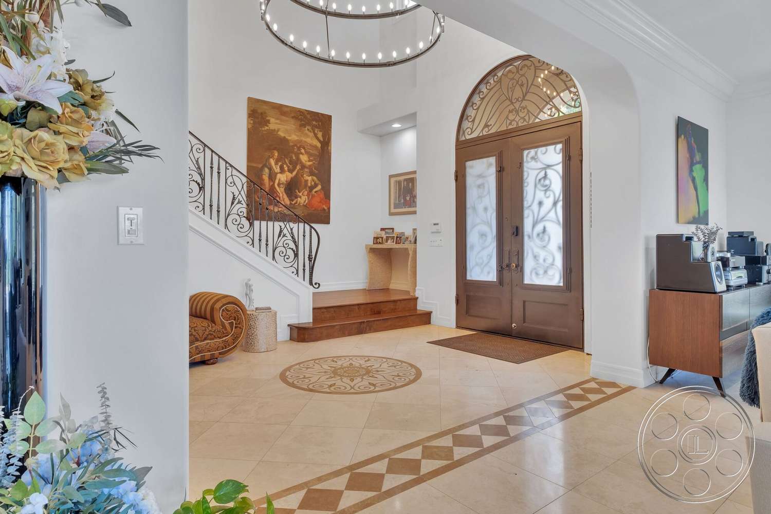 Medit 172 - high ceilings, neutral color palette, recessed lighting, ornate chandelier, textured white painted walls, decorative wall artwork, marble foyer flooring, modern accent furniture, open foyer space, intricate tile inlay design, wood front door with decorative wrought iron, exposed staircase with wooden treads, curved wrought iron railing, flower arrangement in large vase