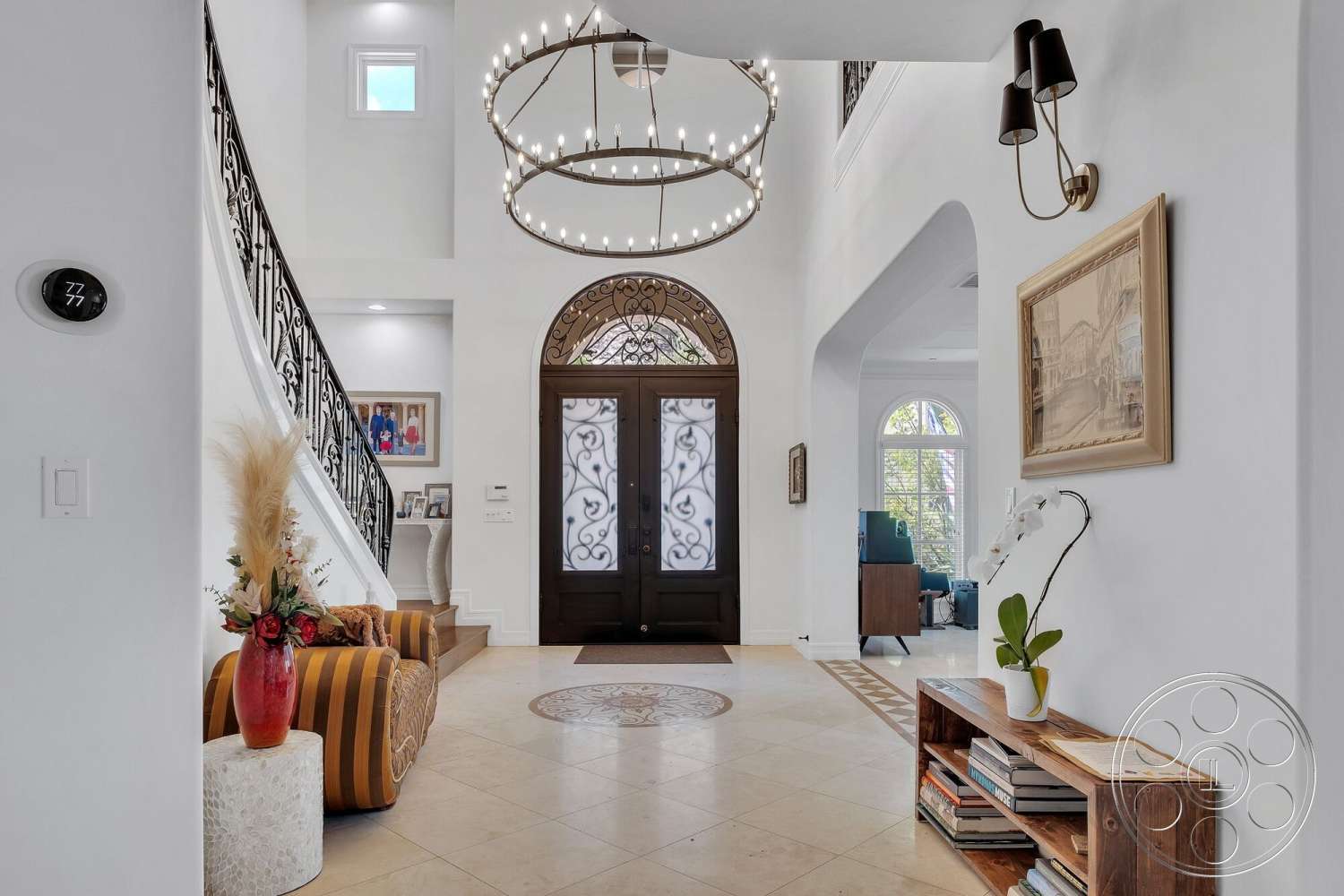 Medit 172 - white painted walls, curved staircase with wrought iron railing, vaulted ceiling with chandelier, ceramic tile foyer flooring, dark wood double front doors with wrought iron details, decorative circular tile inlay, wood console table with books and plant, striped upholstered accent chair, rounded concrete side table with floral arrangement