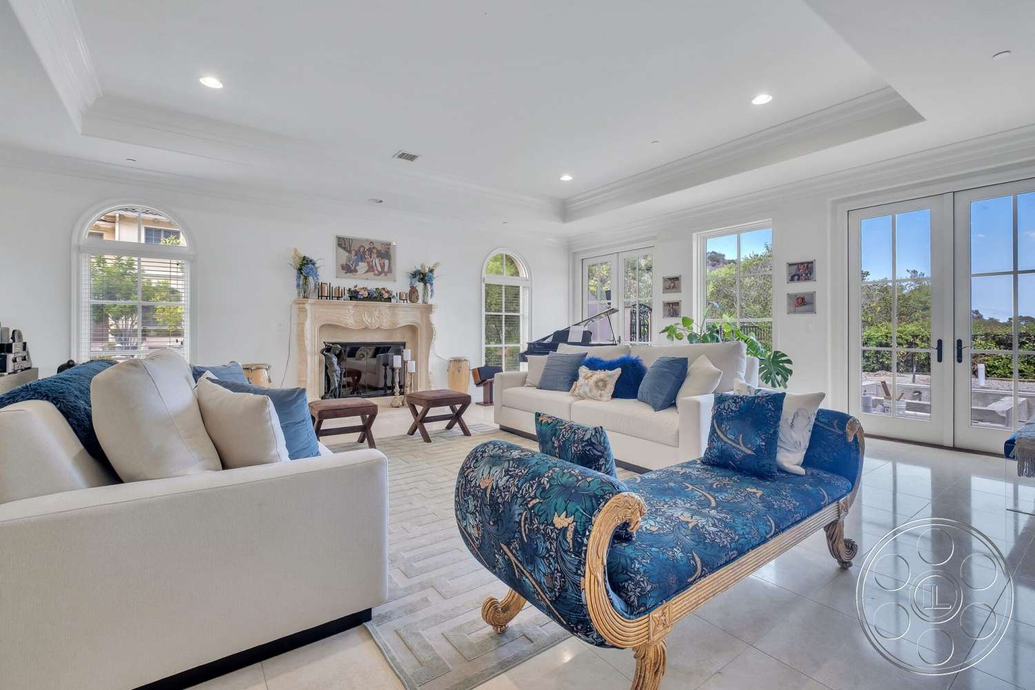 Medit 172 - floor-to-ceiling windows, greenery visible through windows, white painted walls, tile living room flooring, decorative artwork on walls, light-colored fabric sofas, exposed stone fireplace wall, soft white ceiling with crown molding details, blue patterned chaise lounge, arched French doors leading to outdoor area, light wood accent tables