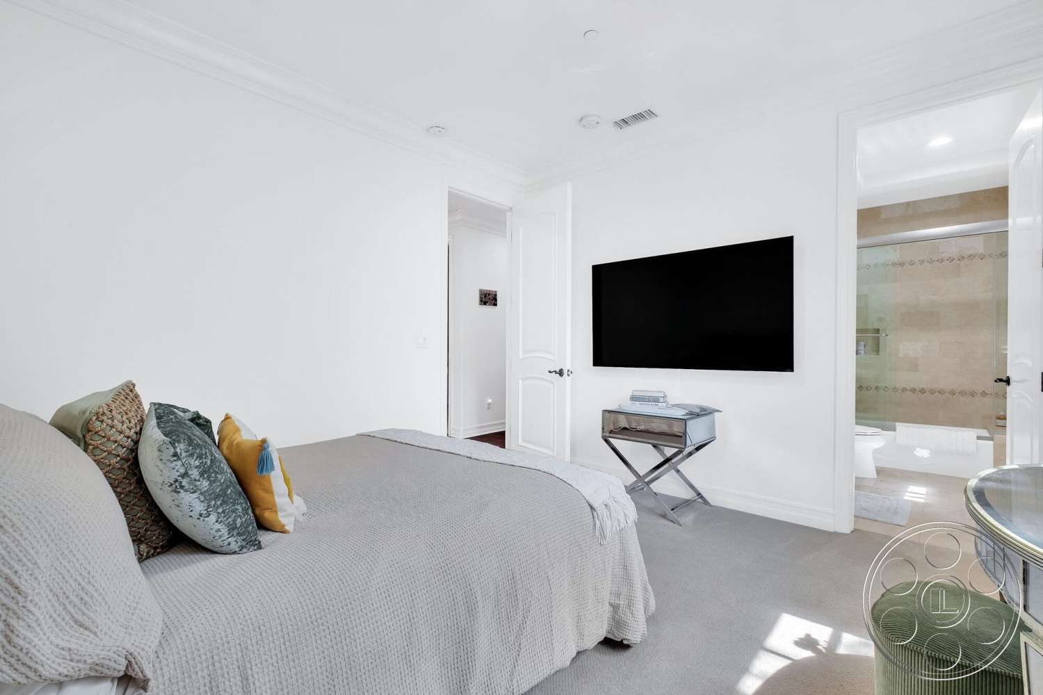 Medit 172 - recessed lighting, textured throw pillows, gray throw blanket, white painted walls, flat-screen wall-mounted television, crown molding details, doorway to bathroom, spacious bedroom layout, plush carpet bedroom flooring, mirror accent side table, light-colored bedspread