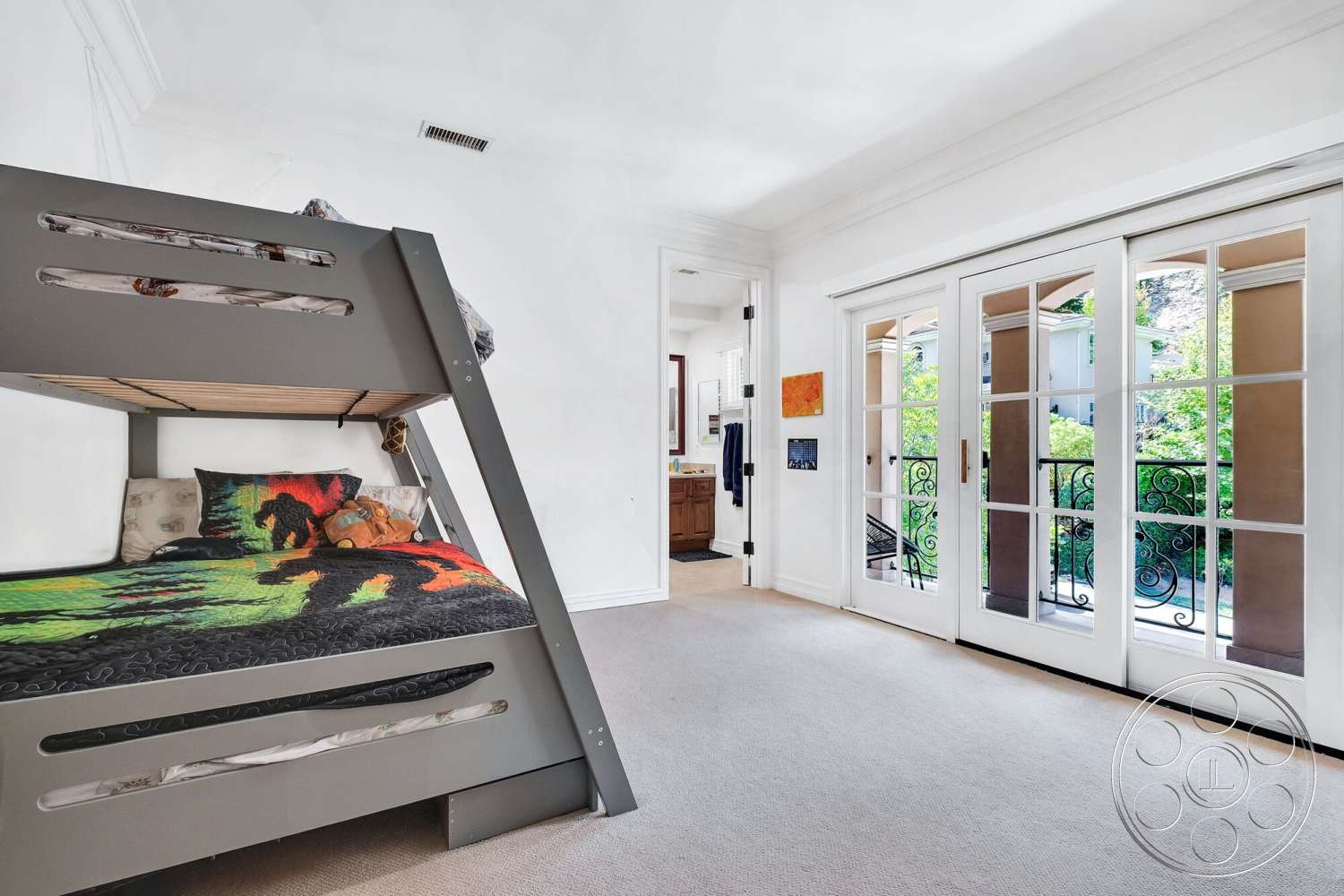 Medit 172 - colorful bedspread, white painted walls, decorative wrought iron balcony railing, neutral carpet flooring, bunk bed with gray metal frame, floor-to-ceiling French doors, natural light from exterior greenery, light crown molding details, warm wooden vanity in adjacent bathroom