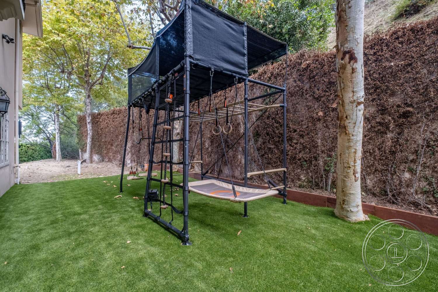 Medit 172 - exterior, mature trees, synthetic grass lawn, concrete border planters, metal playground structure, black fabric shade sail, wooden platform deck, climbing ladder, gymnastic rings, framed swing bench, landscaped hedge backdrop
