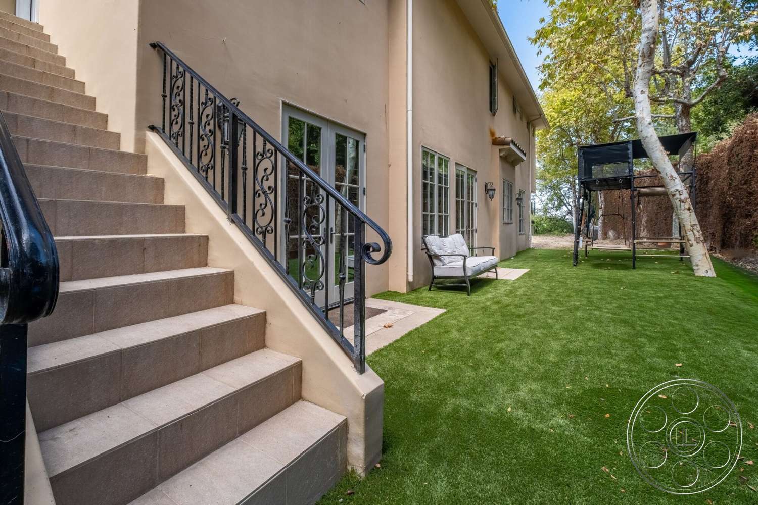 Medit 172 - exterior, concrete steps, modern outdoor seating, stucco wall finish, outdoor patio area, decorative wrought iron railings, landscaped artificial grass lawn, dual-pane windows with grid patterns, tree-lined backyard setting