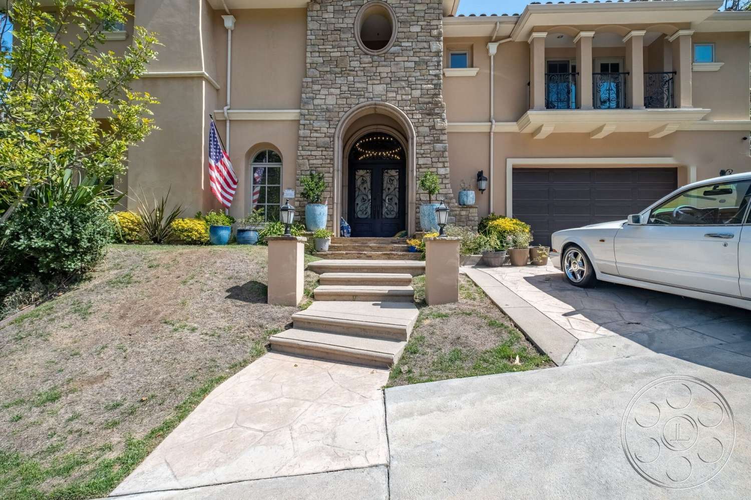 Medit 172 - exterior, potted plants, stucco wall finish, landscaped garden beds, concrete staircase steps, two-story residential home, outdoor lantern fixtures, American flag display, arched entry door, front yard with grass, stone entryway columns, stamped concrete pathway, decorative stone facade, garage door with horizontal panels