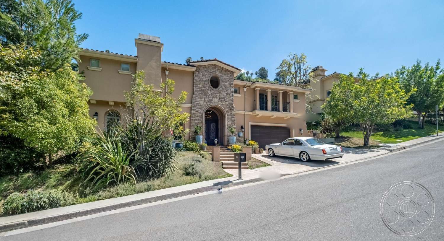 Medit 172 - exterior, flat roof, garage door, stone accent wall, landscaped front yard, window shutters, paved walkway, concrete driveway, arched entryway, decorative front door, hillside location, stucco exterior walls, tree-lined street view, two-story home design, gravel border landscaping, horizontal roof eaves, lawn with ornamental shrubs, attached two-car garage