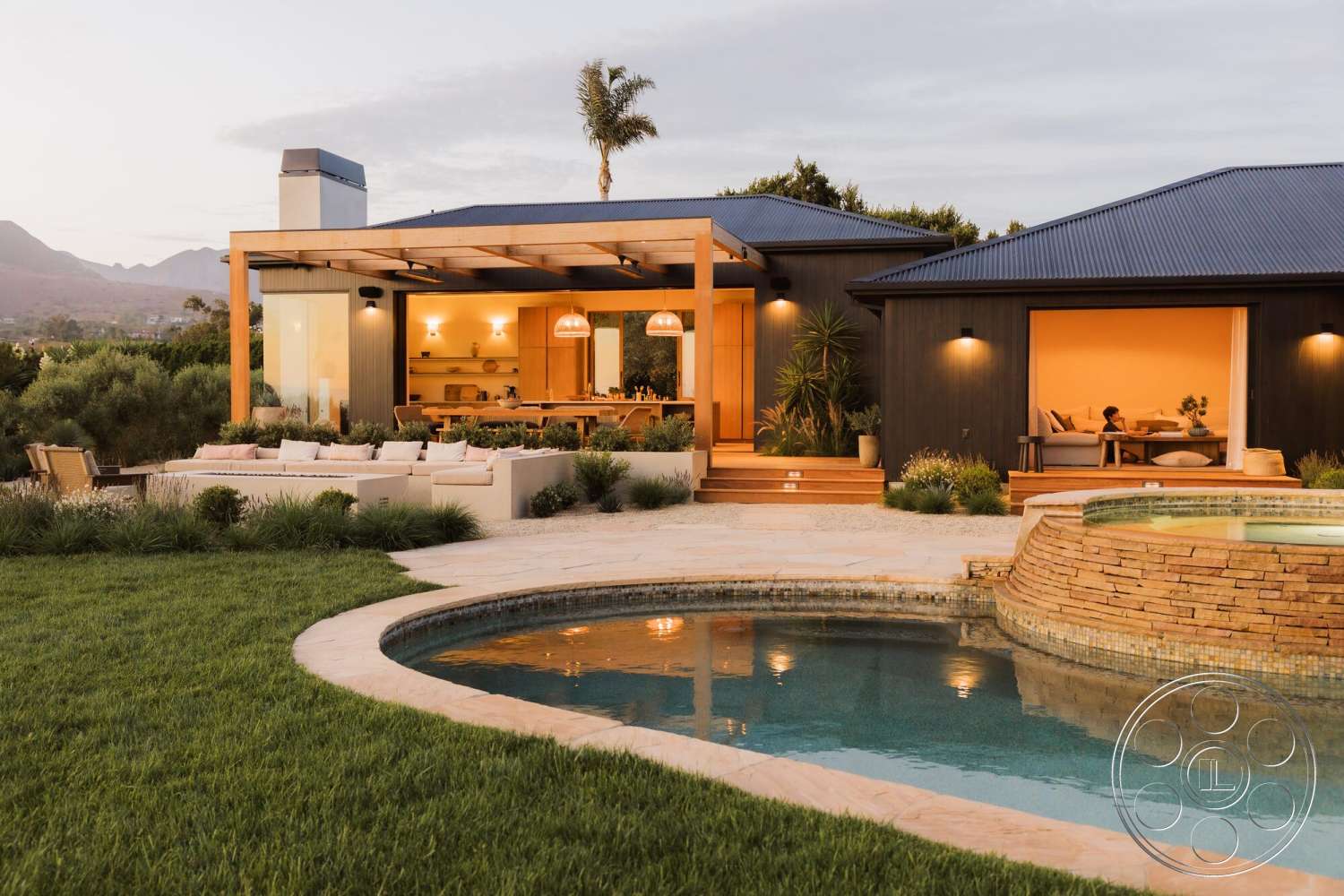 Modern 533 - exterior, landscaped garden, stone accent wall, rectangular swimming pool, floor-to-ceiling windows, lush green grass, mountain view, warm wood paneling, concrete patio flooring, wood beam pergola, open concept kitchen, built-in kitchen cabinets, modern single-story home, luxury outdoor seating area, natural stone pool deck, glass door opening to patio, dusk setting