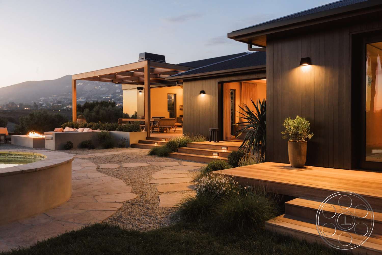 Modern 533 - exterior, glass sliding doors, outdoor seating area, stone pathway, decorative potted plants, planted landscaping, horizontal wood siding, mountain view setting, wooden deck steps, modern pergola structure, curved spa pool, light-colored stone patio flooring, lawn area with ornamental grasses, low concrete wall with fire pit