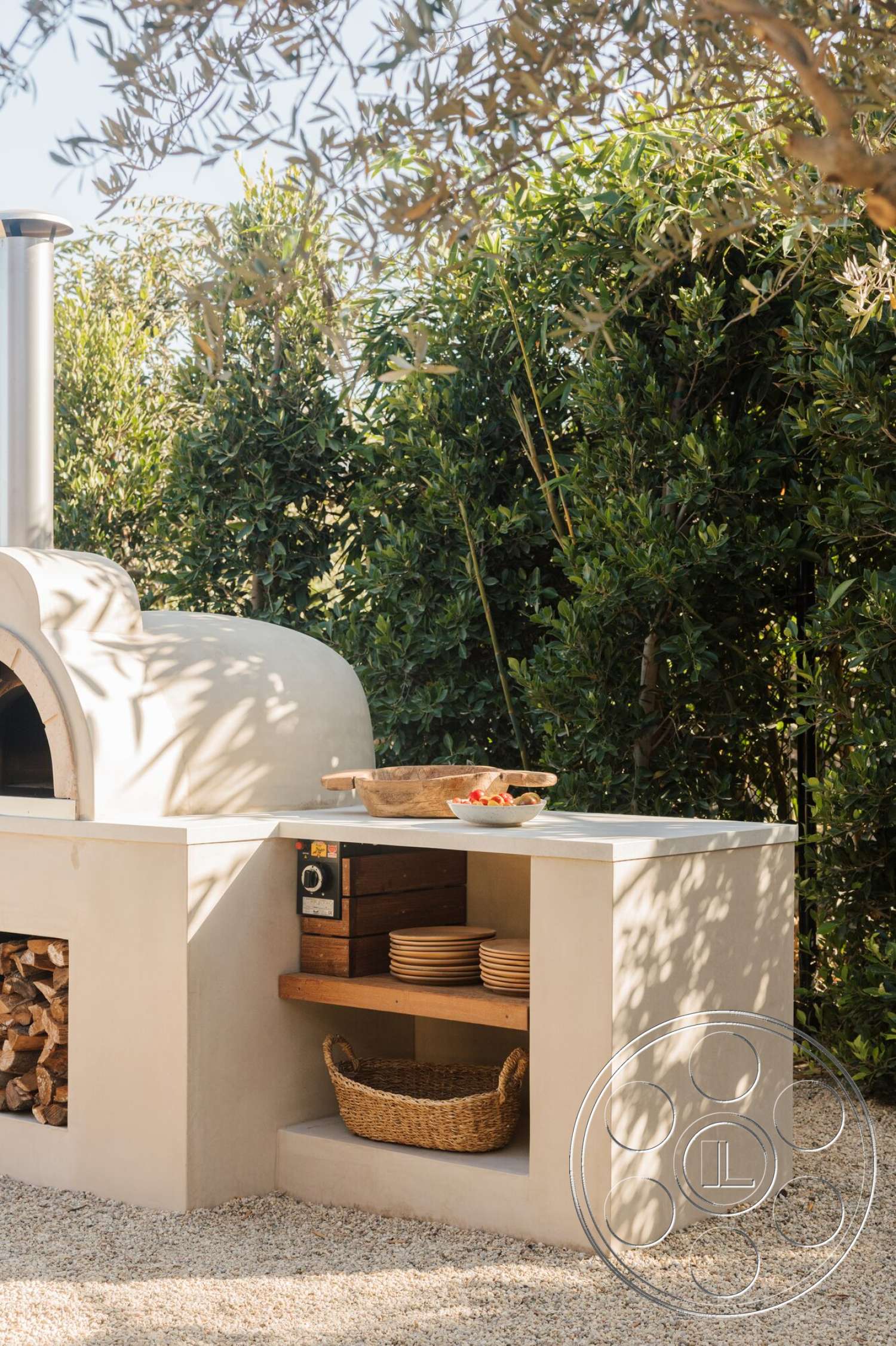 Modern 533 - exterior, lush greenery backdrop, rustic outdoor setting, gravel patio floor, woven basket storage, concrete outdoor kitchen countertop, rounded oven structure, wood storage compartment, wood shelving support