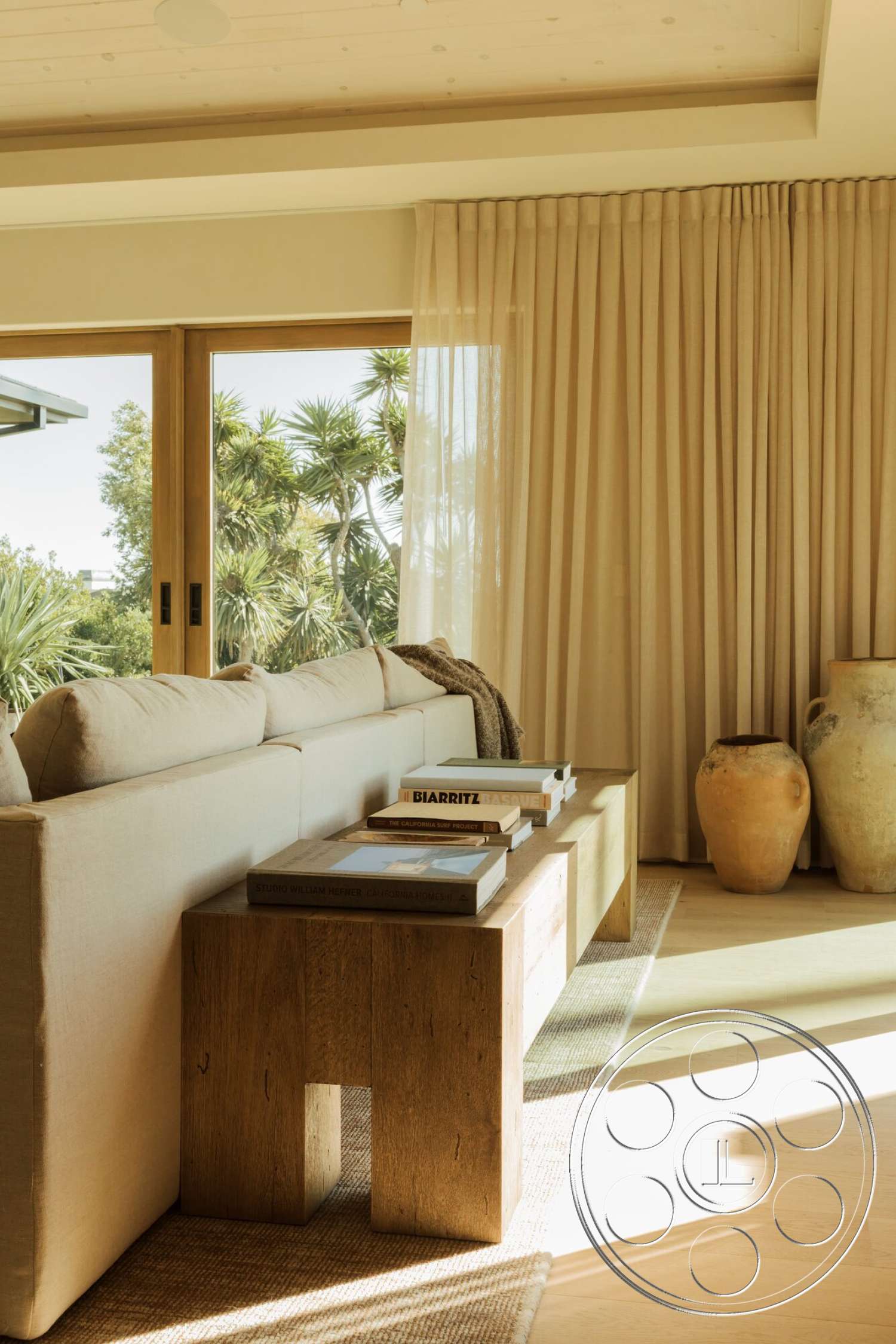 Modern 533 - natural light, wood coffee table, textured area rug, sheer curtains, large glass sliding door, palm tree view, beige fabric sofa, light wood ceiling beams, modern minimalist style, earthy ceramic vases, stacked coffee table books