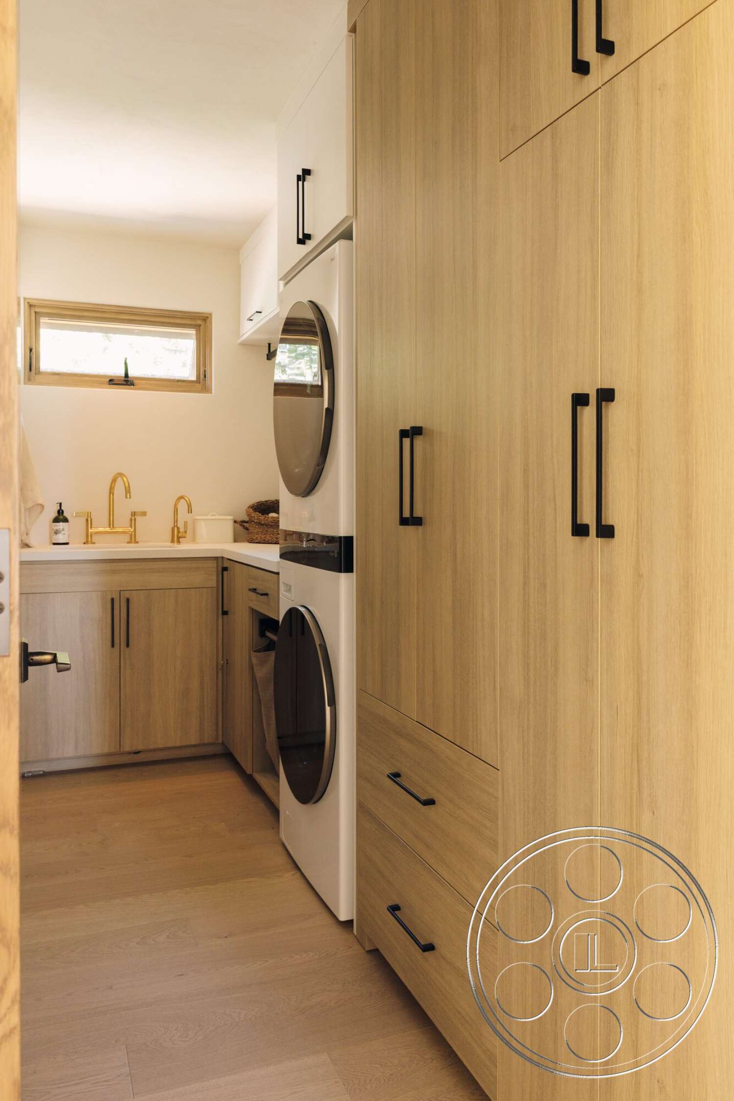 Modern 533 - minimalist design, light hardwood flooring, light wood cabinetry, under-cabinet lighting, gold faucet fixtures, interior laundry room, white front-loading washer, small rectangular window, white front-loading dryer, black handle pulls