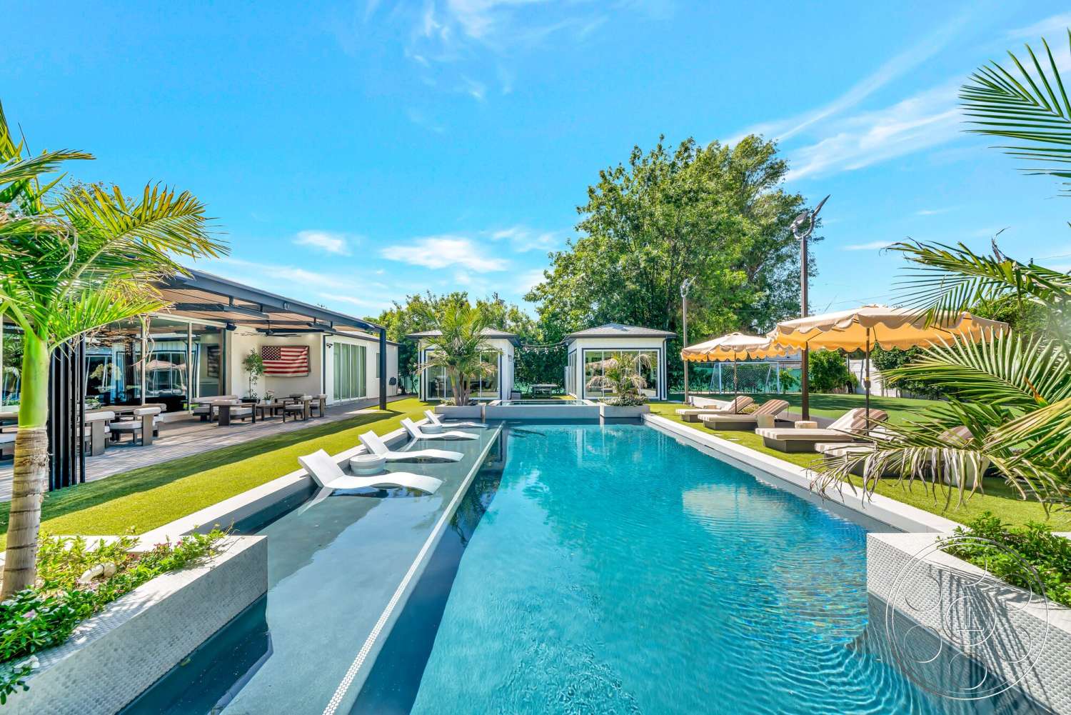 Modern 534 - exterior, palm trees, natural greenery, lounge chairs, rectangular pool, glass patio doors, stone pool deck, landscaped garden area, concrete pool border, wooden patio dining table, synthetic grass lawn, blue sky backdrop, umbrella-covered seating area, modern outdoor cabana, steel outdoor lighting fixtures, flag-themed wall art