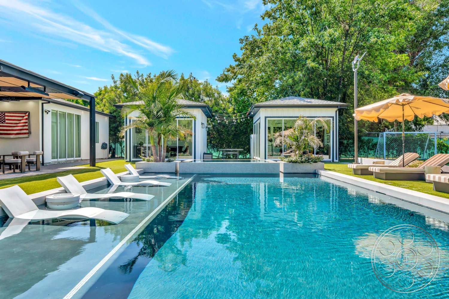 Modern 534 - exterior, glass sliding doors, tropical landscaping, green grass lawn, concrete pool deck, wooden patio furniture, modern outdoor seating, umbrella shade, flagstone patio surface, rectangular infinity pool, glass backyard cabanas