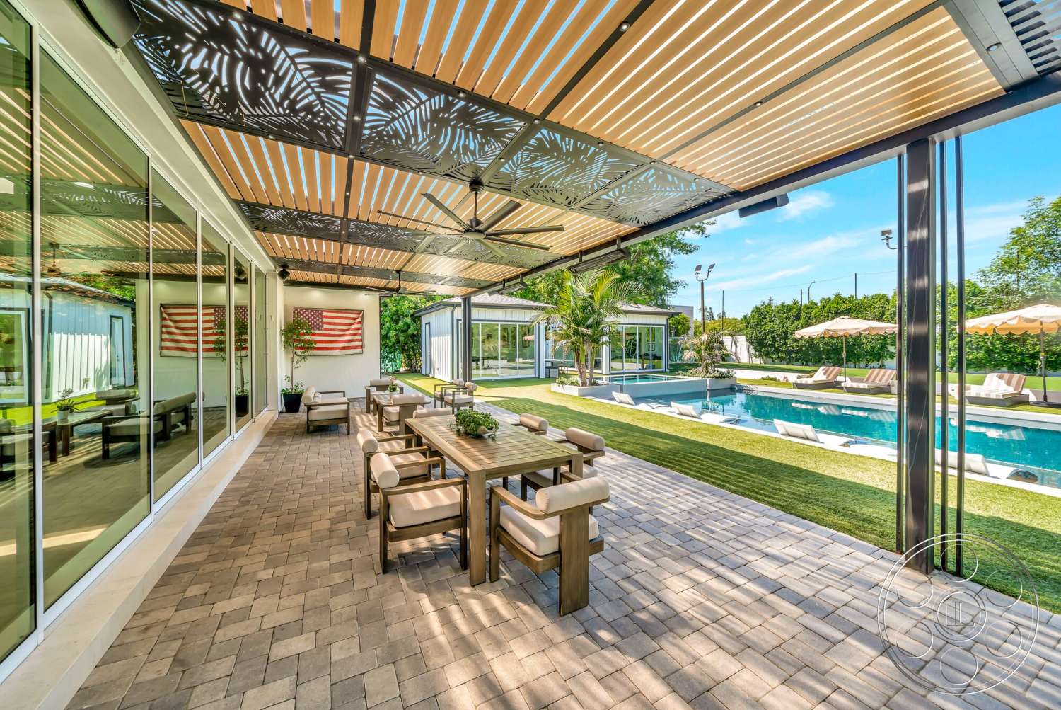 Modern 534 - exterior, rectangular swimming pool, outdoor lounge chairs, floor-to-ceiling glass windows, wooden outdoor dining table, landscaped garden with tropical plants, grassy lawn area, modern outdoor living space, paver stone patio flooring, metal roofed pergola, striped fabric umbrellas, American flag wall art, wooden accents in seating