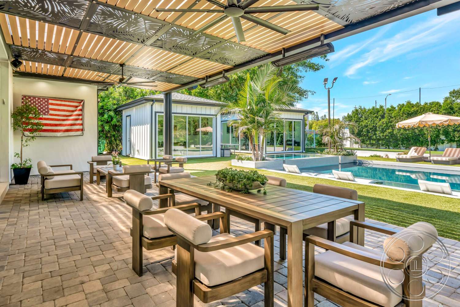 Modern 534 - exterior, upholstered dining chairs, wood dining table, landscaped surroundings, outdoor dining area, rectangular pool, deck chairs, paver patio flooring, lawn with artificial grass, tropical palm plants, pergola with decorative slats, flag wall art, modernist garden design, contemporary shed structure