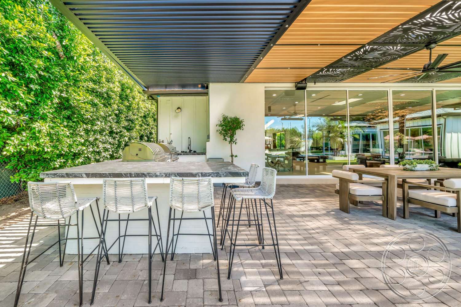 Modern 534 - exterior, wooden dining table, stainless steel grill, marble countertop, metal bar stools, modern architectural style, open-air patio, outdoor kitchen area, floor-to-ceiling glass windows, paver patio flooring, decorative metal ceiling panels, lush green hedge backdrop