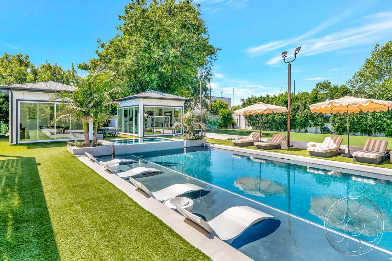 Modern 534 - exterior, modern architectural style, infinity pool, green grass lawn, rectangular pool, outdoor umbrellas, bright blue sky, glass panel walls, palm landscaping, concrete patio flooring, concrete pool border, sunny backyard setting, striped outdoor lounge chairs, dual guest house structures