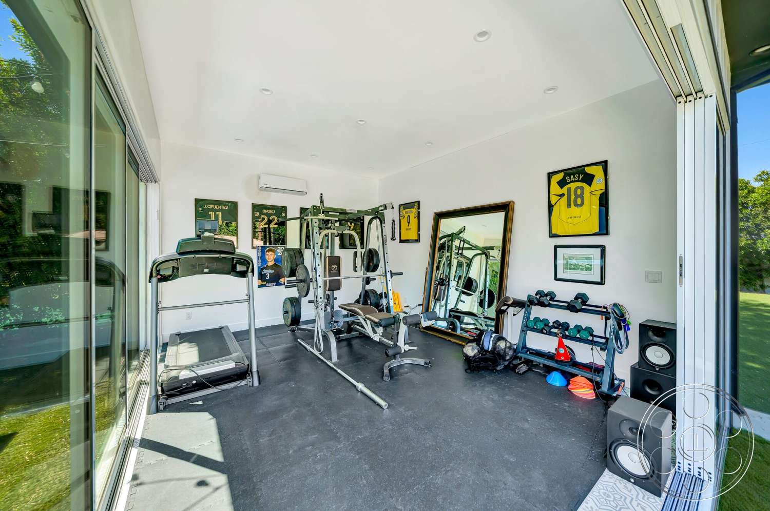 Modern 534 - minimalist decor, open concept layout, natural light from large windows, modern fitness equipment, sliding glass door entrance, interior gym with rubber flooring, fitness room with large mirror, padded exercise bench, metal weightlifting rack, wooden wall-mounted frames, ceiling-mounted air conditioning unit