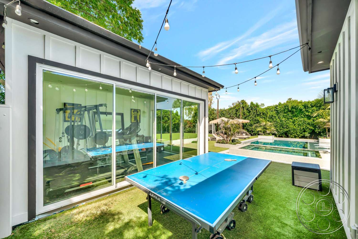 Modern 534 - exterior, flat roof, concrete pool deck, rectangular pool, wooden outdoor furniture, landscaped garden area, artificial grass lawn, white vertical siding, glass wall gym enclosure, outdoor lighting string