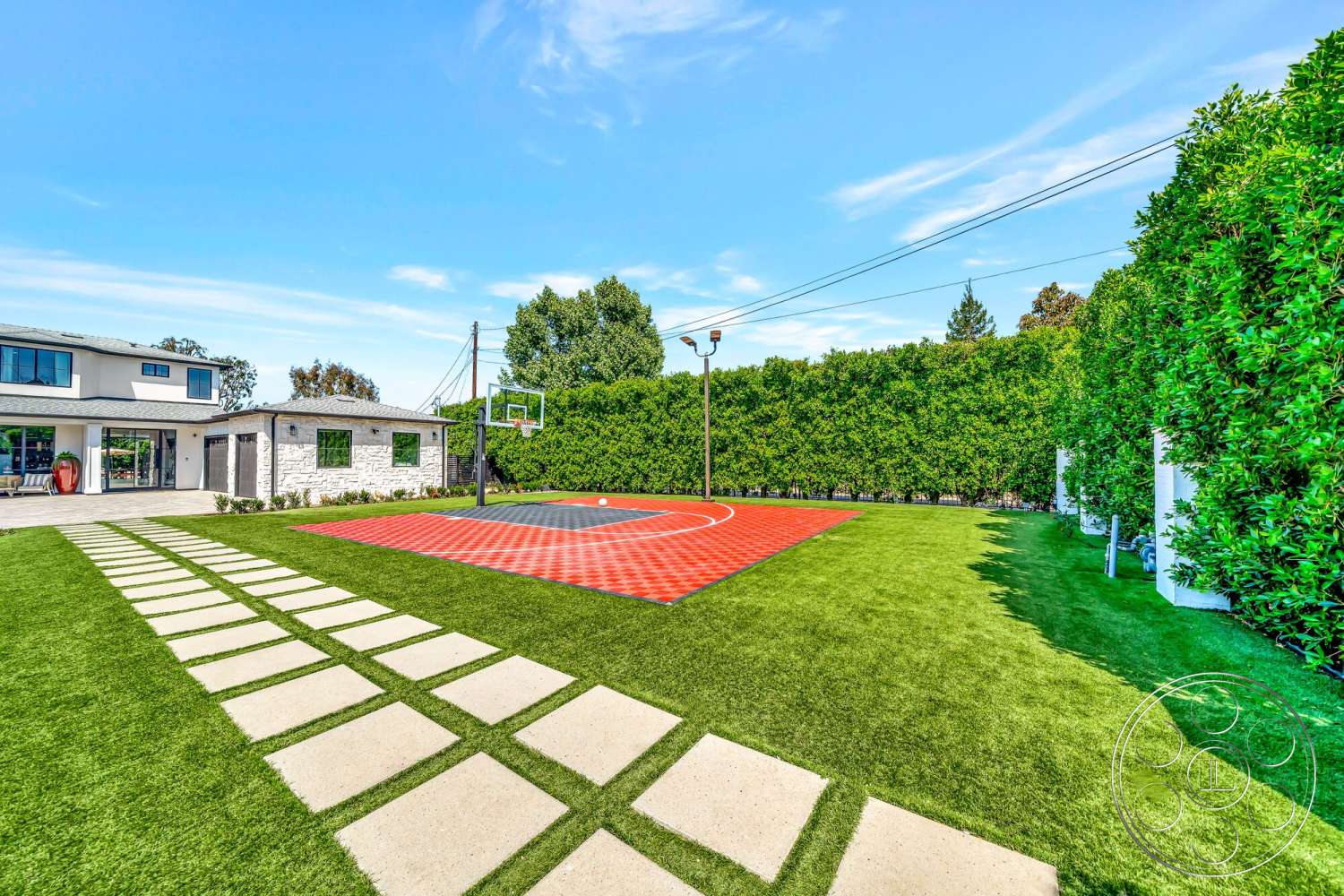 Modern 534 - exterior, natural stone accents, sloped terrain, concrete driveway, stucco walls, outdoor sports area, modern two-story home, sunny outdoor environment, urban backyard setting, synthetic grass lawn, landscaped hedge, red basketball court surface, concrete stepping stones path, rectangular basketball court, patio with outdoor access