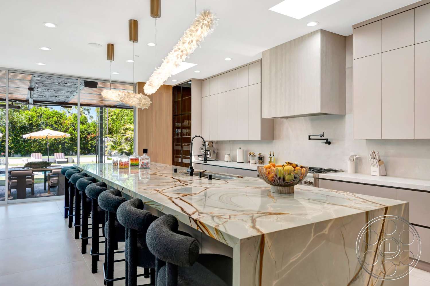 Modern 534 - open floor plan, stainless steel appliances, modern bar stools, pendant lighting fixtures, contemporary architectural style, built-in kitchen cabinets, white cabinetry with minimalist design, marble kitchen island with veining, textured wood paneling on one wall, large glass windows overlooking patio, neutral-colored tile flooring, ceiling skylights, lush outdoor garden view