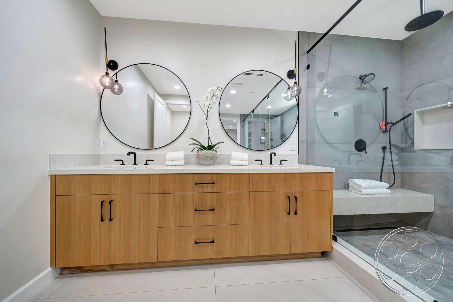Modern 534 - glass shower enclosure, round mirrors, white marble countertop, textured plaster walls, black pendant lighting, large format tile flooring, black faucet fixtures, maple wood bathroom vanity, concrete shower bench, towels on countertop, modern aesthetic bathroom design
