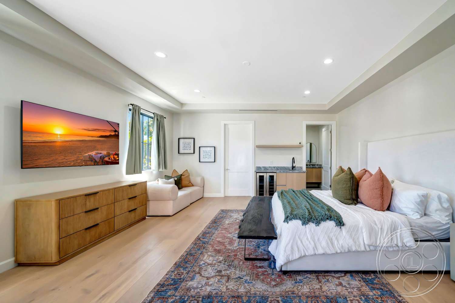Modern 534 - textured throw pillows, decorative rug, light-colored bedding, light wood dresser, hardwood bedroom floors, neutral painted walls, ambient recessed lighting, crown molding details, wall-mounted flat-screen TV, modern upholstered bed, open doorway to bathroom, window with drapery, simple side table, built-in kitchenette with countertop