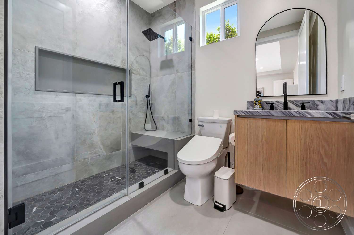 Modern 534 - neutral color palette, glass shower enclosure, white toilet, natural light from window, hexagonal tile flooring, minimalist bathroom design, wood vanity cabinet, marble shower walls, black faucet fixtures, mirrored vanity surface