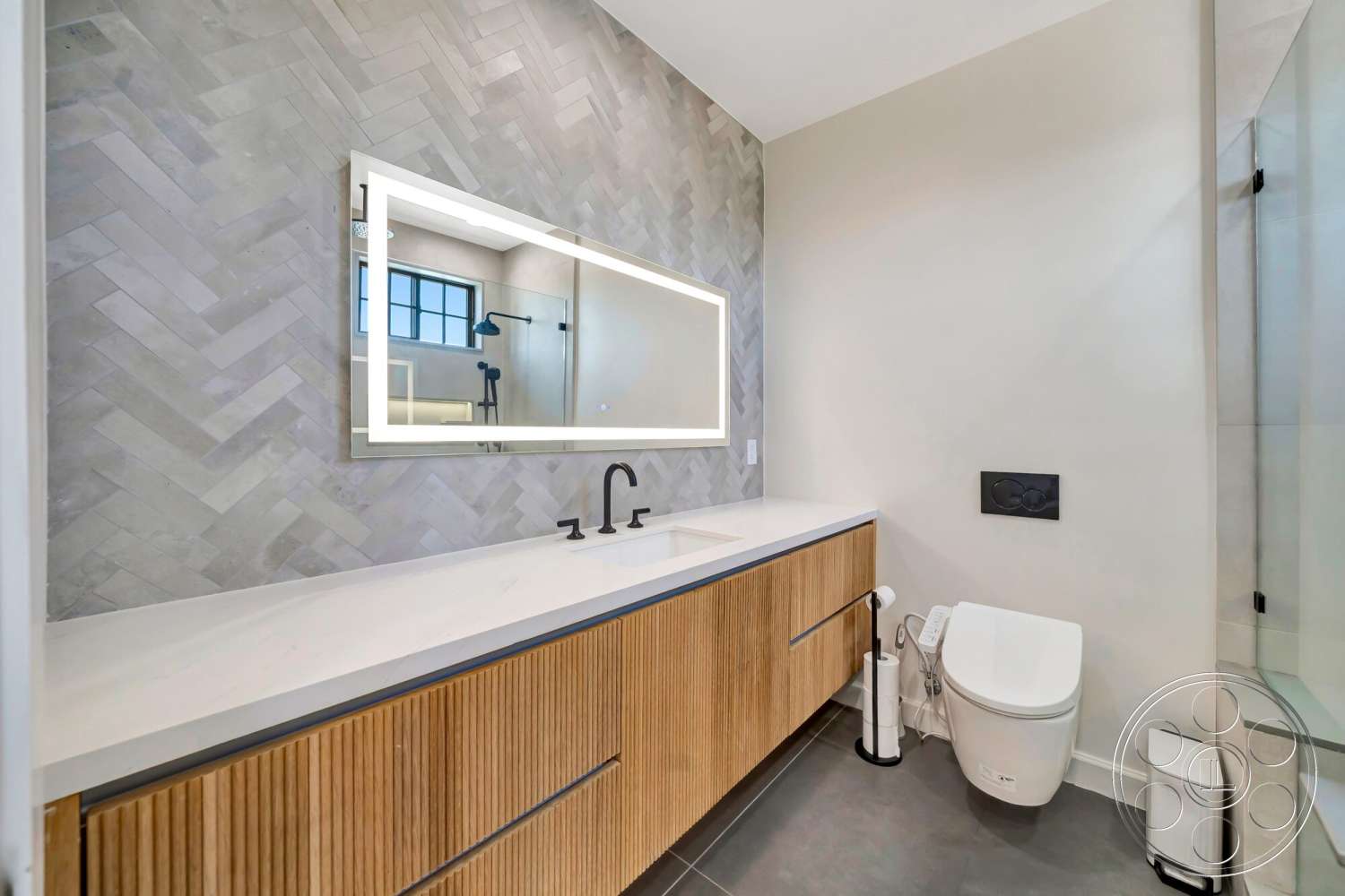 Modern 534 - glass shower enclosure, light wood cabinetry, backlit mirror, white quartz bathroom countertop, minimalist toilet design, black matte faucet fixtures, gray herringbone tile walls, dark tile bathroom flooring