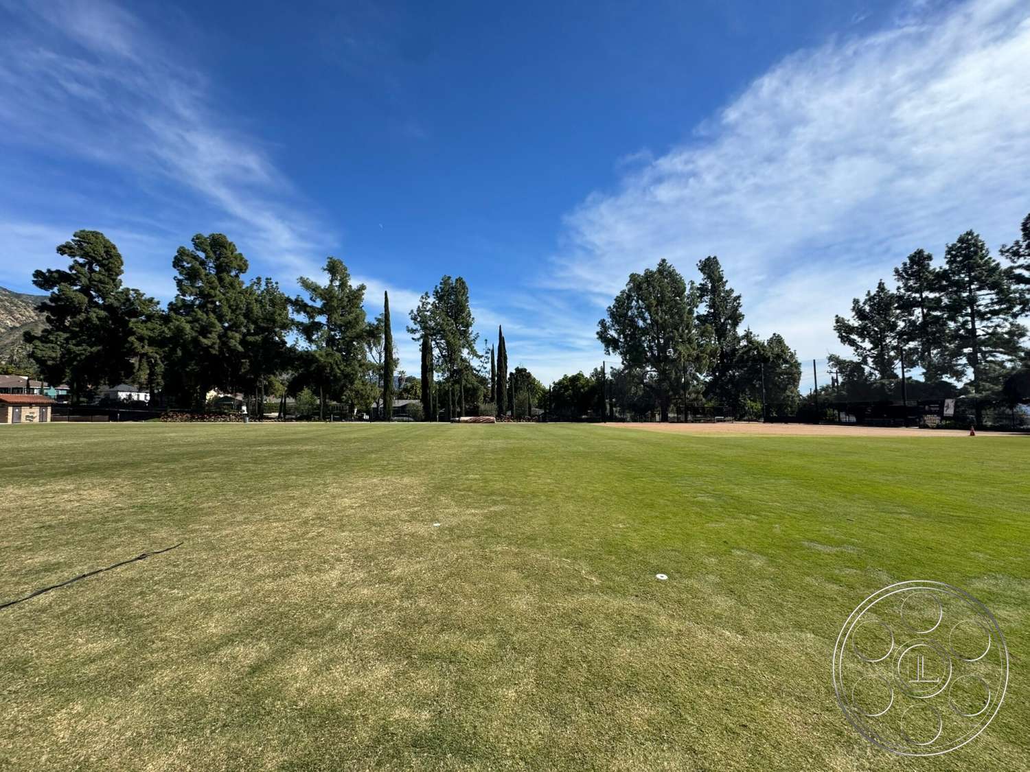 Villa Italy - tall evergreen trees, sports field, natural outdoor setting, landscaped lawn area, distant mountain backdrop, open grassy field, well-maintained grass, blue sky with clouds, park environment, shaded areas from trees, community recreational area