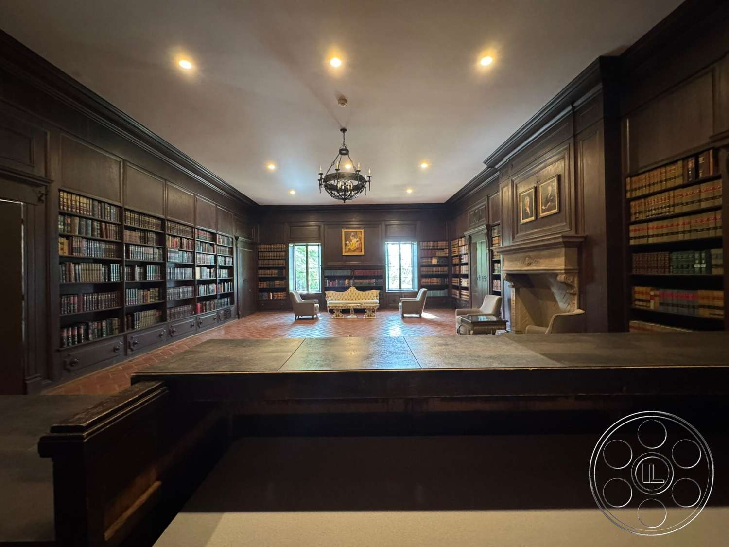 Villa Italy - stone fireplace, wooden coffee table, framed artwork, dark wood paneling, double-hung windows, built-in bookshelves, exposed brick fireplace surround, coffered ceiling details, traditional chandelier lighting, terra cotta tile flooring, library room, classic armchairs, luxurious reading nook
