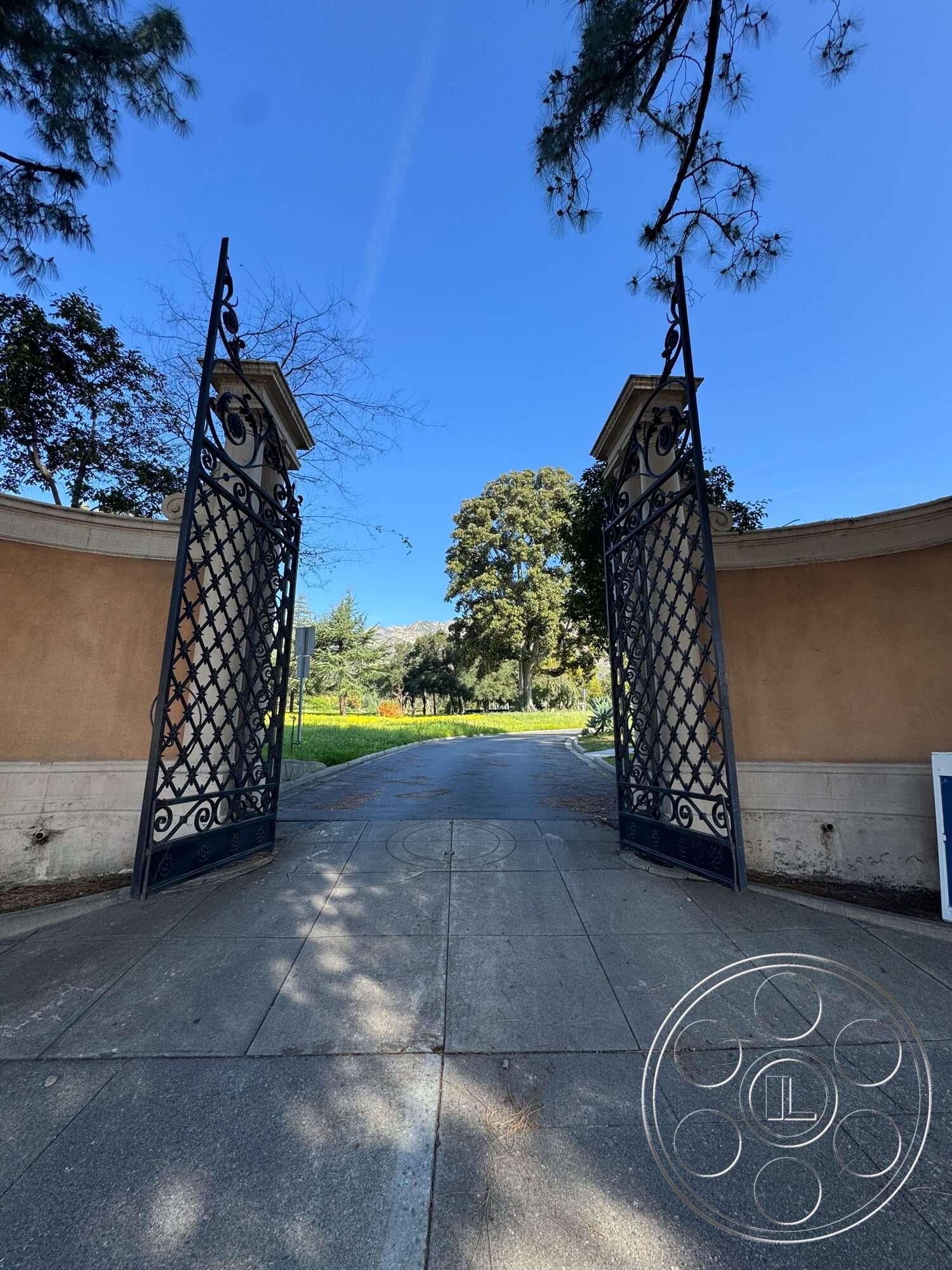 Villa Italy - landscaped garden, lush greenery, concrete driveway, exterior entrance, blue sky backdrop, tree-lined pathway, tan stucco wall, ornate wrought iron gates, circular stone driveway pattern
