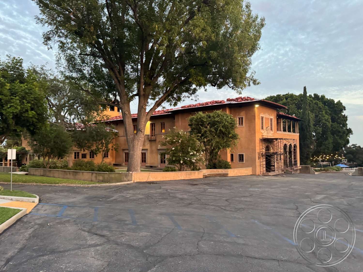 Villa Italy - exterior, exposed wooden beams, large trees, red tile roof, stucco walls, arched windows, Mediterranean architectural style, paved parking lot, two-story home, landscaped garden area, decorative wrought iron details