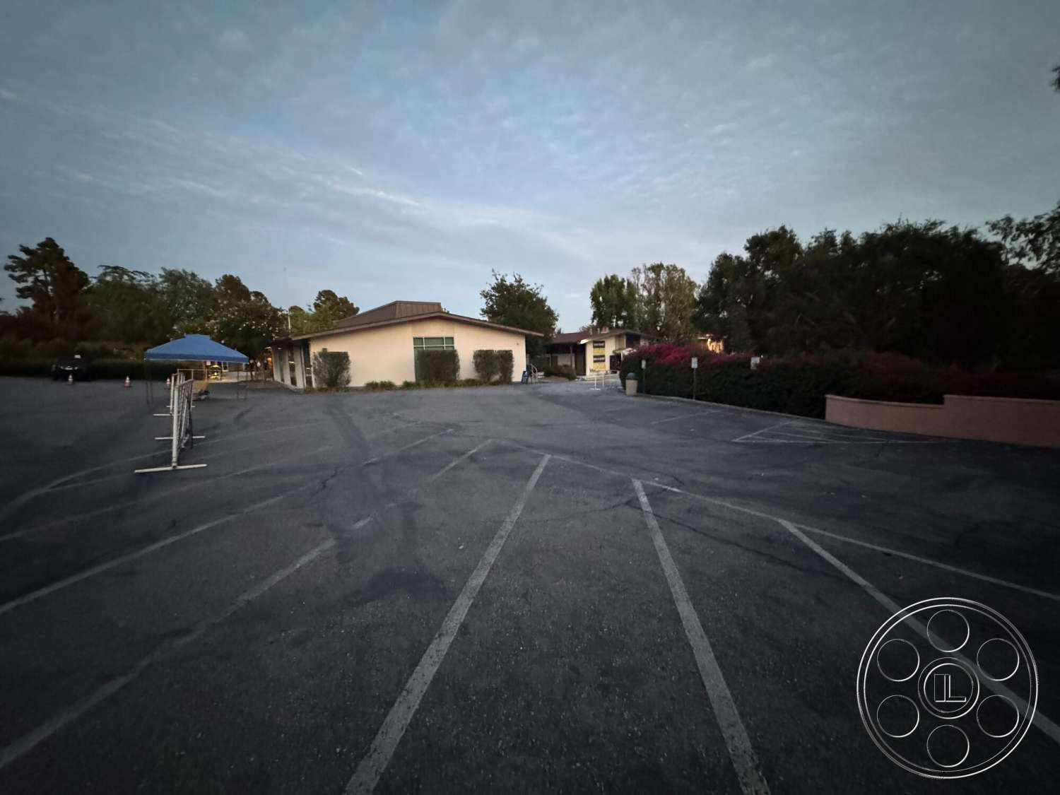 Villa Italy - large windows, surrounding trees, gabled roof, asphalt parking lot, single-story building, landscaped garden area, exterior lighting fixtures, concrete flower bed border, metal roof overhang, fenced entry area, evening dusk lighting