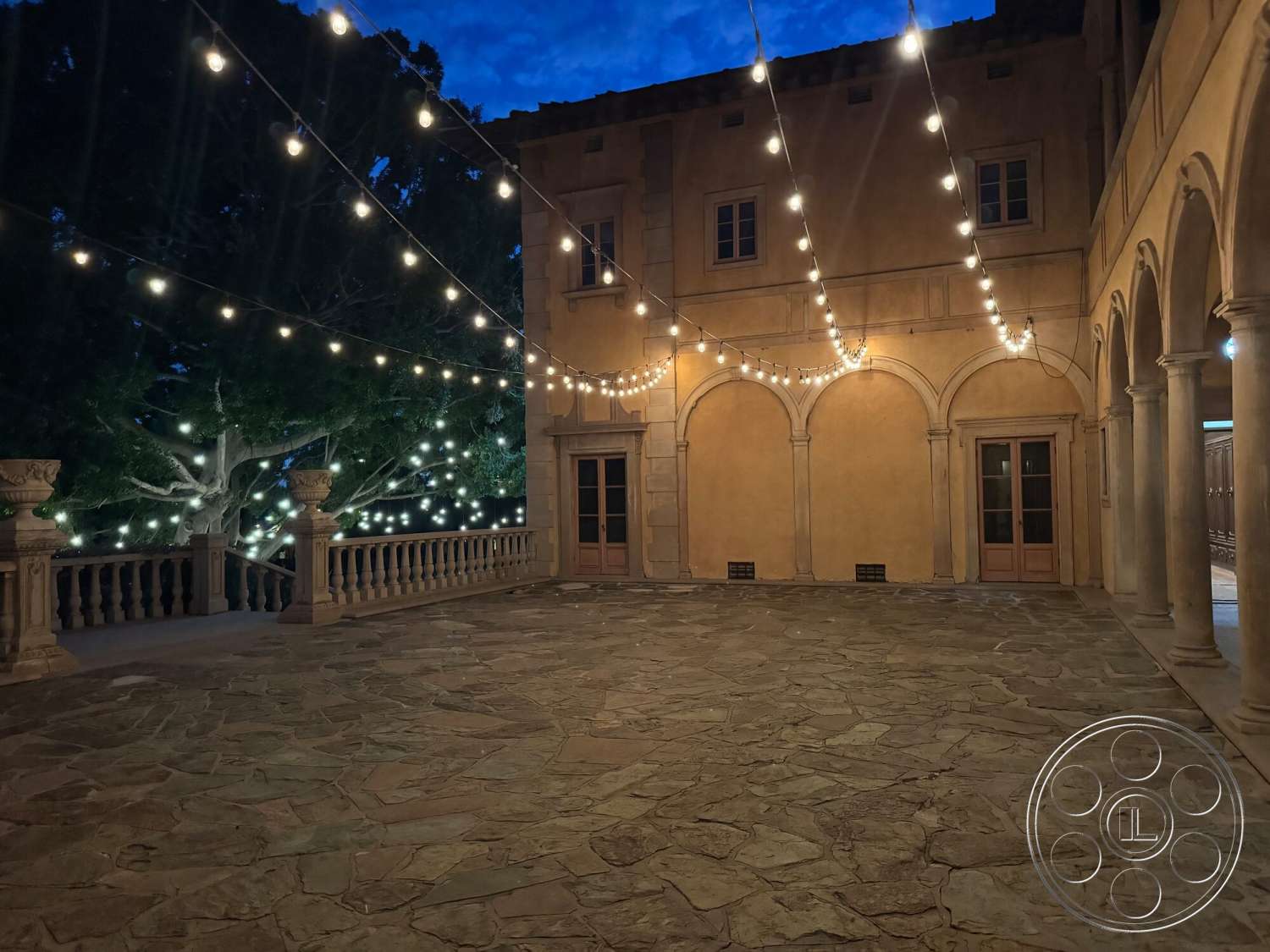 Villa Italy - exterior, outdoor dining area, string lights, landscape lighting, arched doorways, columned porch, natural stone patio flooring, stucco wall finishes, dark wood double doors, tree canopy backdrop, Italian architectural style