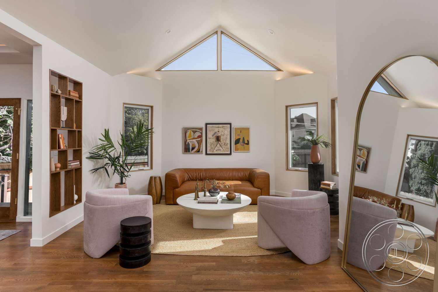 Craftsman 65 - modern coffee table, dark wood flooring, framed windows, tan leather sofa, white painted walls, decorative indoor plants, light-colored area rug, wooden bookshelf with open shelving, angular ceiling with skylight, rounded pink upholstered armchairs, gallery-style wall art, mirrored full-length standing mirror