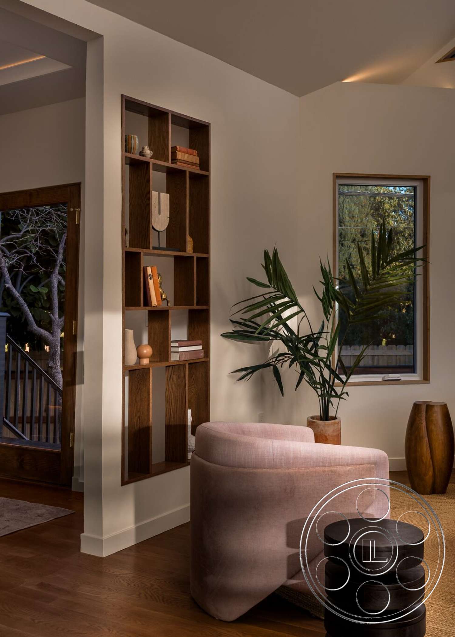 Craftsman 65 - minimalist decor, hardwood flooring, natural lighting, textured beige area rug, dark wood accent table, white painted walls, large potted plant, large window with wooden frame, exposed wood shelving unit, curved pink upholstered lounge chair